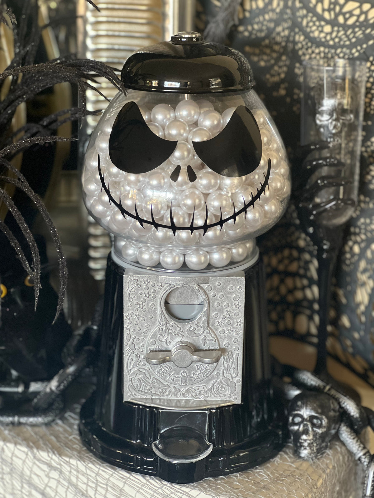 A skull-themed gumball machine prop decorated with white faux pearls, black plastic details, and a silver ornate front panel, styled in a Halloween setting with spiderwebs and black decorative props.