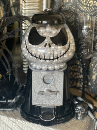 A skull-themed gumball machine prop decorated with white faux pearls, black plastic details, and a silver ornate front panel, styled in a Halloween setting with spiderwebs and black decorative props.