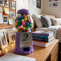 A purple vintage-style gumball machine filled with colorful gumballs sits on a wooden desk in a bright, sunlit bedroom or dorm room. Books, framed photos, a bed with pillows, and posters are visible in the background.