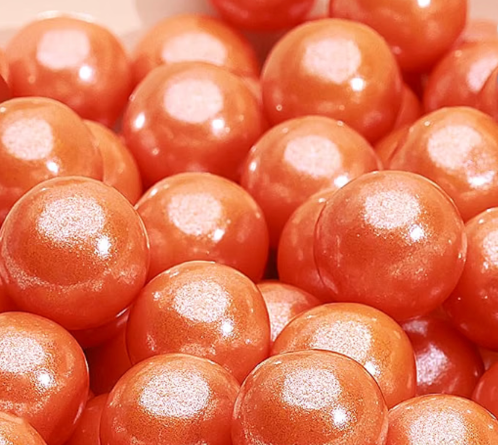 A close-up pile of glossy, metallic orange-red spherical beads or candies with a pearlescent finish.