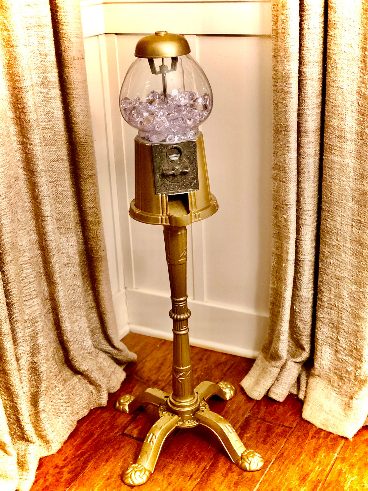 A gold pedestal gumball machine filled with fake diamonds, placed in a room with wood floors. 
